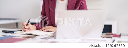 Asian adult designer professional drafting creative project plans at bright office desk with laptop and material samples 129267220