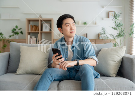 Smiling handsome young Asian man sitting on cozy couch 129268071