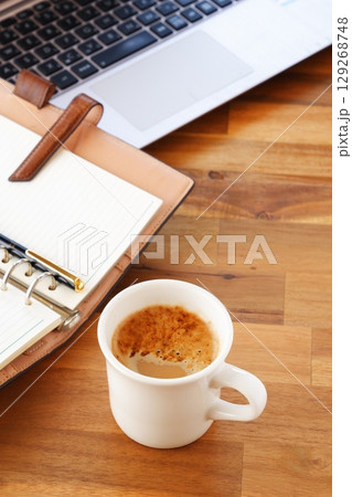 リング式のシステム手帳を広げ、コーヒー（カフェ・ラテ）を飲みながらパソコンでデスクワーク中のイメージ 129268748