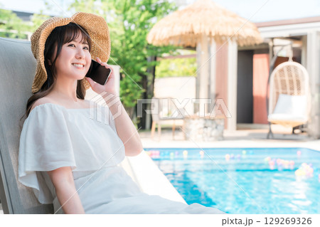 プルサイドのリクライニングチェアに座りスマホで通話する若いアジア人女性（予約,検索,ルームサービス） 129269326