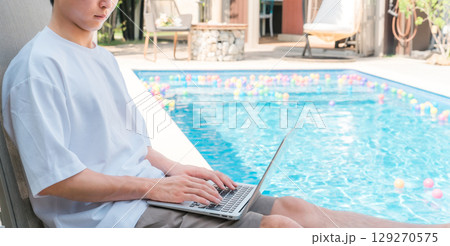 プールサイドのリクライニングチェアに座りパソコンを操作する若いアジア人男性 129270575