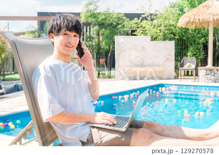 プールサイドのリクライニングチェアに座りスマホで通話する若いアジア人男性（予約,ルームサービス） 129270578