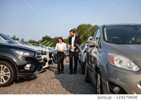 A male car dealer assisting a female customer as they walk through a car lot, possibly discussing vehicle options. A male car dealer assisting a female customer as they walk through a car lot, possibly discussing vehicle options. 129270606