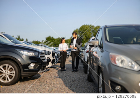 A male car dealer assisting a female customer in a car lot, discussing vehicle options and features. A male car dealer assisting a female customer in a car lot, discussing vehicle options and features. 129270608