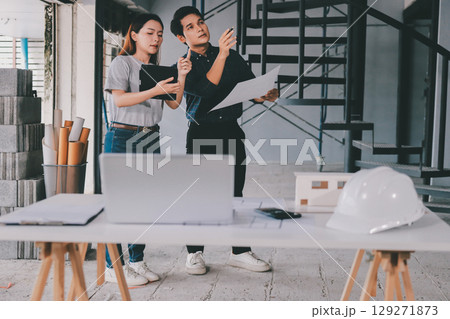 Construction manager and engineer dressed in orange work vests and hard helmets explore construction documentation on the building site near the steel frames 129271873