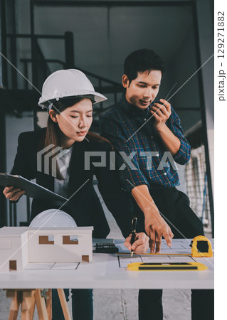 Construction manager and engineer dressed in orange work vests and hard helmets explore construction documentation on the building site near the steel frames Construction manager and engineer dressed in orange work vests and hard helmets explore construction documentation on the building site near the steel frames 129271882