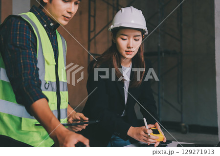 Construction manager and engineer dressed in orange work vests and hard helmets explore construction documentation on the building site near the steel frames 129271913