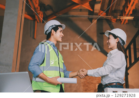 Construction manager and engineer dressed in orange work vests and hard helmets explore construction documentation on the building site near the steel frames Construction manager and engineer dressed in orange work vests and hard helmets explore construction documentation on the building site near the steel frames 129271928