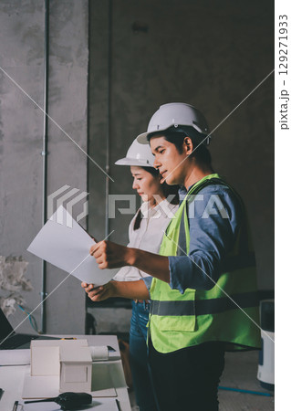 Construction manager and engineer dressed in orange work vests and hard helmets explore construction documentation on the building site near the steel frames 129271933