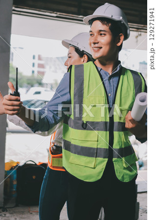 Construction manager and engineer dressed in orange work vests and hard helmets explore construction documentation on the building site near the steel frames Construction manager and engineer dressed in orange work vests and hard helmets explore construction documentation on the building site near the steel frames 129271941