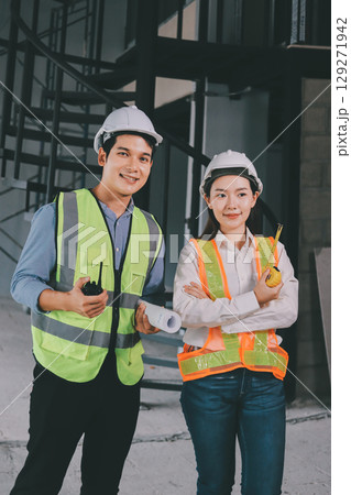 Construction manager and engineer dressed in orange work vests and hard helmets explore construction documentation on the building site near the steel frames Construction manager and engineer dressed in orange work vests and hard helmets explore construction documentation on the building site near the steel frames 129271942