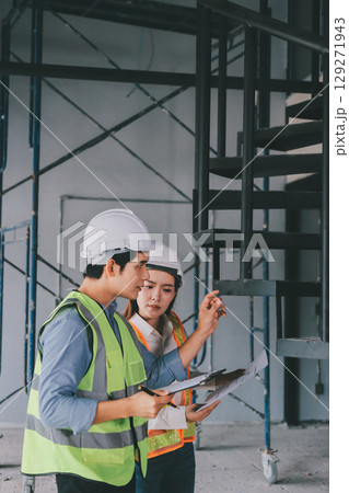 Construction manager and engineer dressed in orange work vests and hard helmets explore construction documentation on the building site near the steel frames Construction manager and engineer dressed in orange work vests and hard helmets explore construction documentation on the building site near the steel frames 129271943
