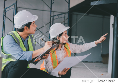 Construction manager and engineer dressed in orange work vests and hard helmets explore construction documentation on the building site near the steel frames Construction manager and engineer dressed in orange work vests and hard helmets explore construction documentation on the building site near the steel frames 129271947