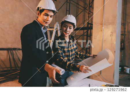 Construction manager and engineer dressed in orange work vests and hard helmets explore construction documentation on the building site near the steel frames Construction manager and engineer dressed in orange work vests and hard helmets explore construction documentation on the building site near the steel frames 129271962
