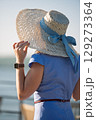 Woman with brown long hair and straw hat in summer dress walking near a lake 129273364