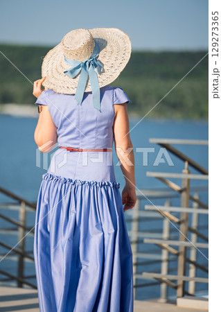 Woman with brown long hair and straw hat in summer dress walking near a lake 129273365