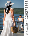 Woman with brown long hair and straw hat and bag in summer dress walking near a lake 129273368