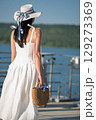Woman with brown long hair and straw hat and bag in summer dress walking near a lake 129273369