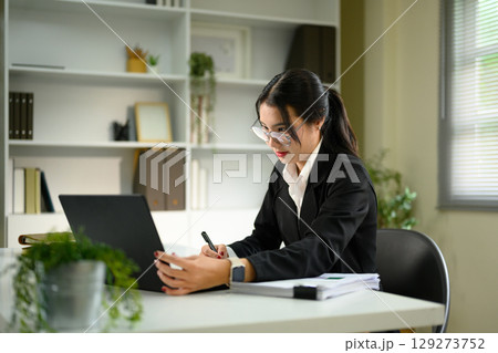 Focused businesswoman working on a laptop while taking notes at a modern desk 129273752
