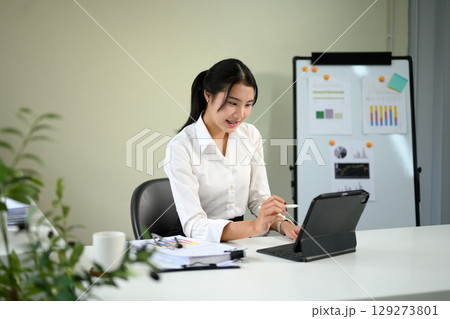Smiling asian employee working on digital tablet at a modern office desk Smiling asian employee working on digital tablet at a modern office desk 129273801