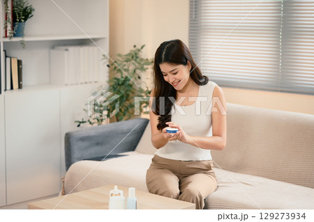 Smiling woman applying moisturizer on her arm for soft skin in a bright living room Smiling woman applying moisturizer on her arm for soft skin in a bright living room 129273934