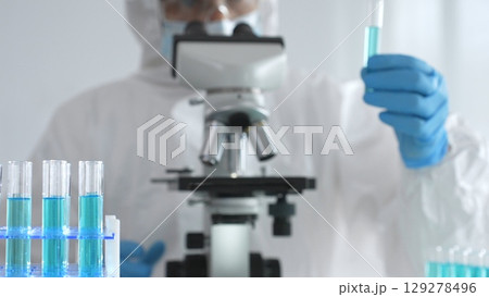 Medical scientist in protective suit and gloves using microscope while analyzing samples, conducting research in laboratory. Close-up of test tubes with blue liquid Medical scientist in protective suit and gloves using microscope while analyzing samples, conducting research in laboratory. Close-up of test tubes with blue liquid 129278496