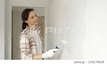 Middle aged woman wearing protective gloves painting white wall with paint roller, updating interior and giving room clean, modern look during home renovation 129278626