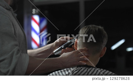 Skilled barber trimming client's hair with electric razor, creating precise contemporary hairstyle inside sleek urban barbershop setting 129278628
