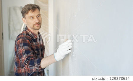 Construction worker man wearing gloves and holding a putty knife is applying plaster on a white wall, performing home renovation and improvement 129278634
