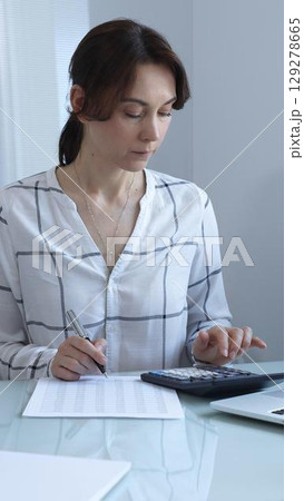 A professional woman calculates finances, checks documents, and accurately and carefully tracks business expenses at a glass table. Audit and taxes in business. 129278665