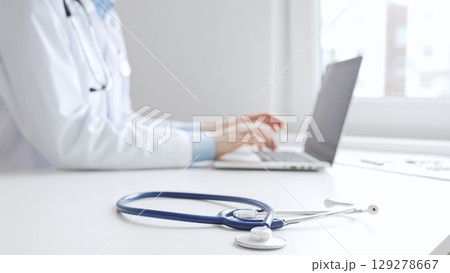 Closeup of blue stethoscope is lying on the table near female doctor working on laptop, highlighting digital tools transforming healthcare diagnostics and patient documentation. Medicine concept 129278667