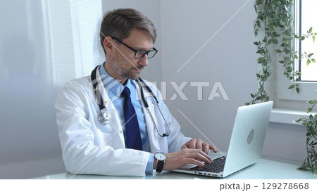 Male doctor wearing a lab coat and stethoscope, sitting at a desk and typing on a laptop computer in a modern medical office, focused on patient care and consultation. Medicine and health care 129278668