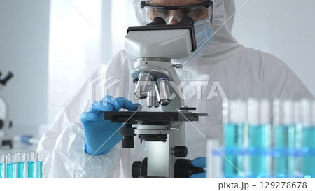 A male doctor in a protective suit and gloves uses a microscope while analyzing samples, conducting research in a laboratory. Close-up of test tubes with blue liquid 129278678