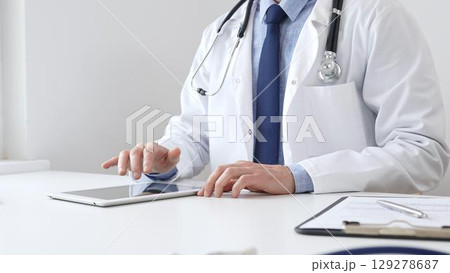 Doctor man wearing white medical coat and stethoscope using digital tablet while sitting at desk in medical office, with medical chart and pen in the foreground. Medicine concept Doctor man wearing white medical coat and stethoscope using digital tablet while sitting at desk in medical office, with medical chart and pen in the foreground. Medicine concept 129278687