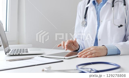 Female doctor wearing white medical coat and stethoscope using digital tablet with laptop and medical records on desk in medical office, showcasing modern healthcare technology. Medicine and helthcare Female doctor wearing white medical coat and stethoscope using digital tablet with laptop and medical records on desk in medical office, showcasing modern healthcare technology. Medicine and helthcare 129278808