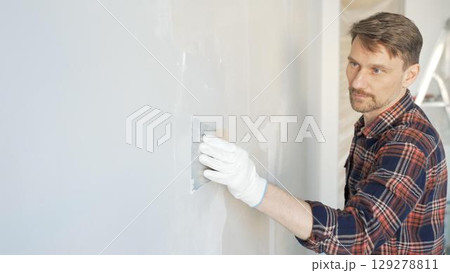 Construction worker man wearing gloves and holding a putty knife is applying plaster on a white wall, performing home renovation and improvement Construction worker man wearing gloves and holding a putty knife is applying plaster on a white wall, performing home renovation and improvement 129278811