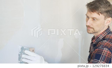 Male construction worker spreading plaster on white wall, wearing protective gloves during home renovation project with putty knife 129278834