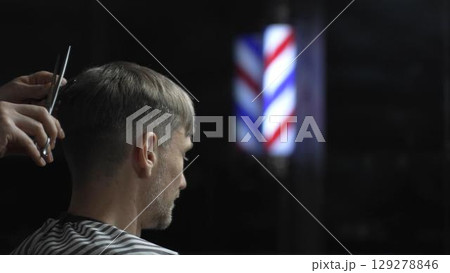Professional barber grooming male client's hair with scissors, vintage barber pole framing traditional barbershop setting Professional barber grooming male client's hair with scissors, vintage barber pole framing traditional barbershop setting 129278846