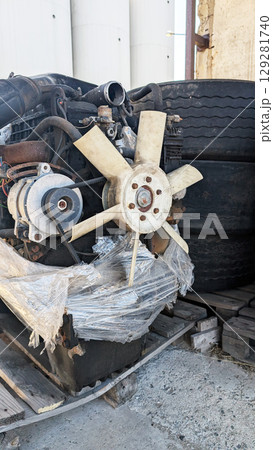 Engine cooling fan assembly with attached components, resting on wooden pallets beside large tires, showcasing industrial machinery and mechanical parts in a storage environment Engine cooling fan assembly with attached components, resting on wooden pallets beside large tires, showcasing industrial machinery and mechanical parts in a storage environment 129281740