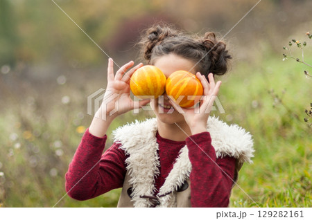 Playful Smiling girl sitting in grassy autumn field holding small pumpkins over her eyes, with a basket of apples beside her, celebrating Thanksgiving outdoors Autumn Harvest. Playful Smiling girl sitting in grassy autumn field holding small pumpkins over her eyes, with a basket of apples beside her, celebrating Thanksgiving outdoors Autumn Harvest. 129282161