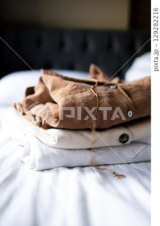 Neatly folded brown, beige, and white clothes on bed with necklace 129282216