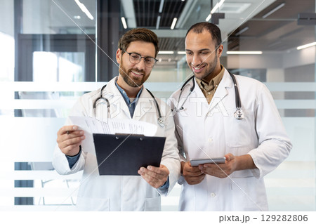 Two doctors, in white coats with stethoscopes, review paperwork and a tablet in a modern office setting. They appear focused and collaborative. 129282806