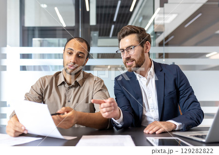 Two diverse businessmen collaborate, reviewing documents at a modern office desk, discussing and analyzing data for a project. Two diverse businessmen collaborate, reviewing documents at a modern office desk, discussing and analyzing data for a project. 129282822