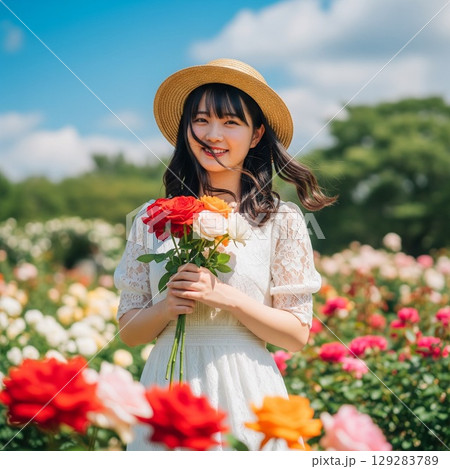 色とりどりの薔薇の花畑で麦わら帽子を被り微笑む女性の夏のポートレート　風に揺れるロングヘアとリボン 129283789