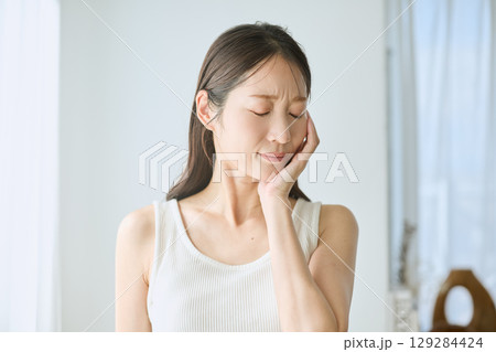 歯の痛みに苦しむ女性・頬を押さえるポーズ・白いタンクトップの若い女性 歯の痛みに苦しむ女性・頬を押さえるポーズ・白いタンクトップの若い女性 129284424