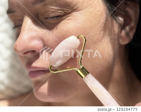 Mature woman lying on cosmetology table during professional facial massage with roller. Anti aging skin care in beauty clinic. Wrinkles, skin care, beauty and wellness concept Mature woman lying on cosmetology table during professional facial massage with roller. Anti aging skin care in beauty clinic. Wrinkles, skin care, beauty and wellness concept 129284775
