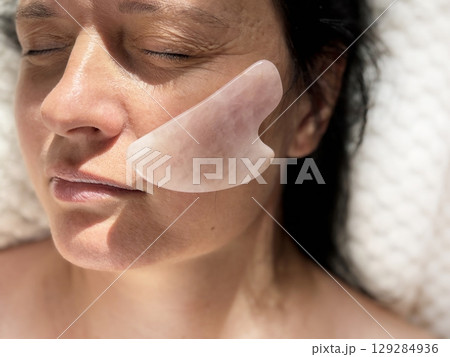 Mature woman lying on cosmetology table during professional facial massage with natural scraper. Anti aging skin care in beauty clinic. Wrinkles, skin care, beauty and wellness concept Mature woman lying on cosmetology table during professional facial massage with natural scraper. Anti aging skin care in beauty clinic. Wrinkles, skin care, beauty and wellness concept 129284936