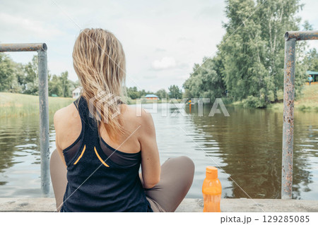 close up woman sitting cross-legged on wooden dock over calm river, wearing sportswear, back view, summer greenery and countryside houses, meditation, morning ritual, nature therapy 129285085