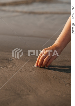 Child's hand touching wet sand at the beach during sunset in a serene coastal setting Child's hand touching wet sand at the beach during sunset in a serene coastal setting 129287655