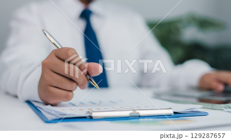 Professional in suit analyzing financial charts and reports with pen, representing business planning, data analysis, accounting, and strategic consultation in corporate office setting. Professional in suit analyzing financial charts and reports with pen, representing business planning, data analysis, accounting, and strategic consultation in corporate office setting. 129287657